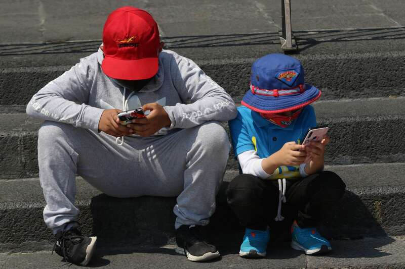 Un par de niños usando dispositivos celulares. Dos niños con gorra y cubrebocas utilizando teléfonos celulares en unos escalones.